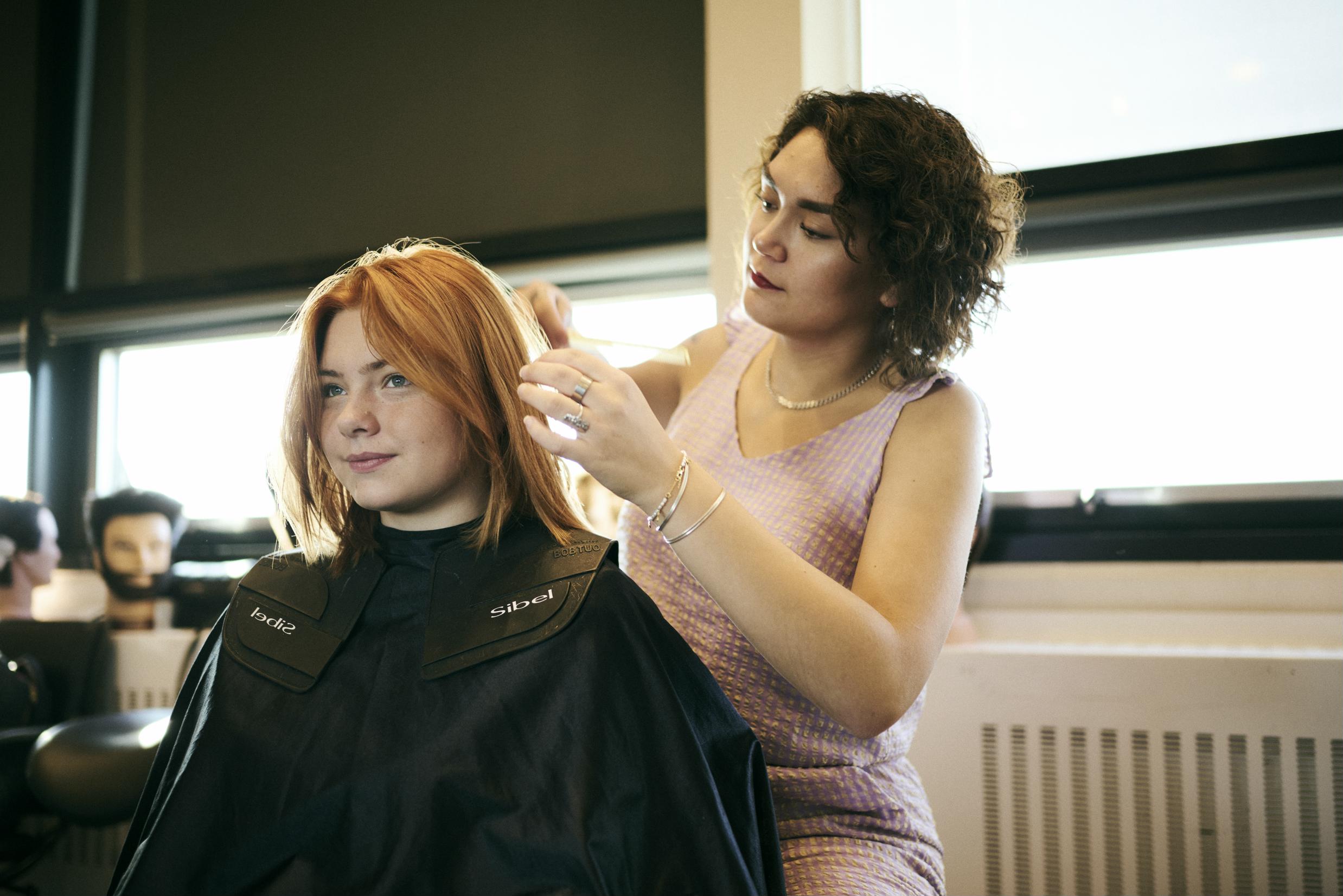 En pige sidder model og får ordnet håret af en frisørelev på Hansenberg.