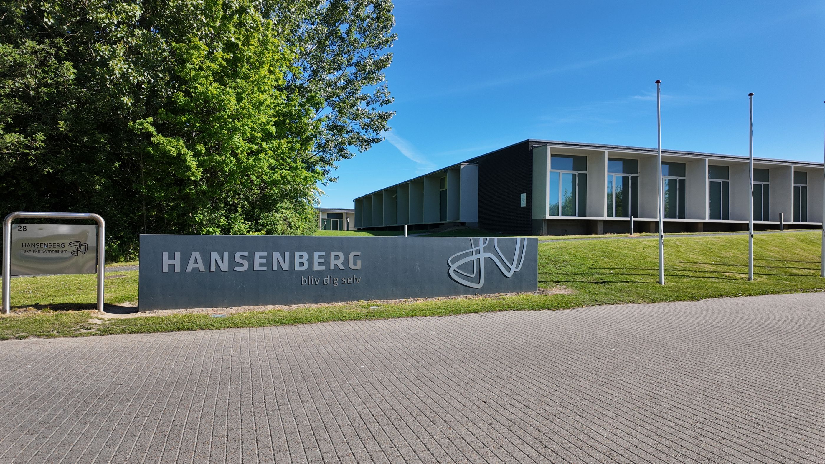 Indkørslen til HANSENBERG på Skovvangen 28 en solskinsdag i maj 2026. Man kan se et udsnit af skolehjemmet bag stenskiltet.