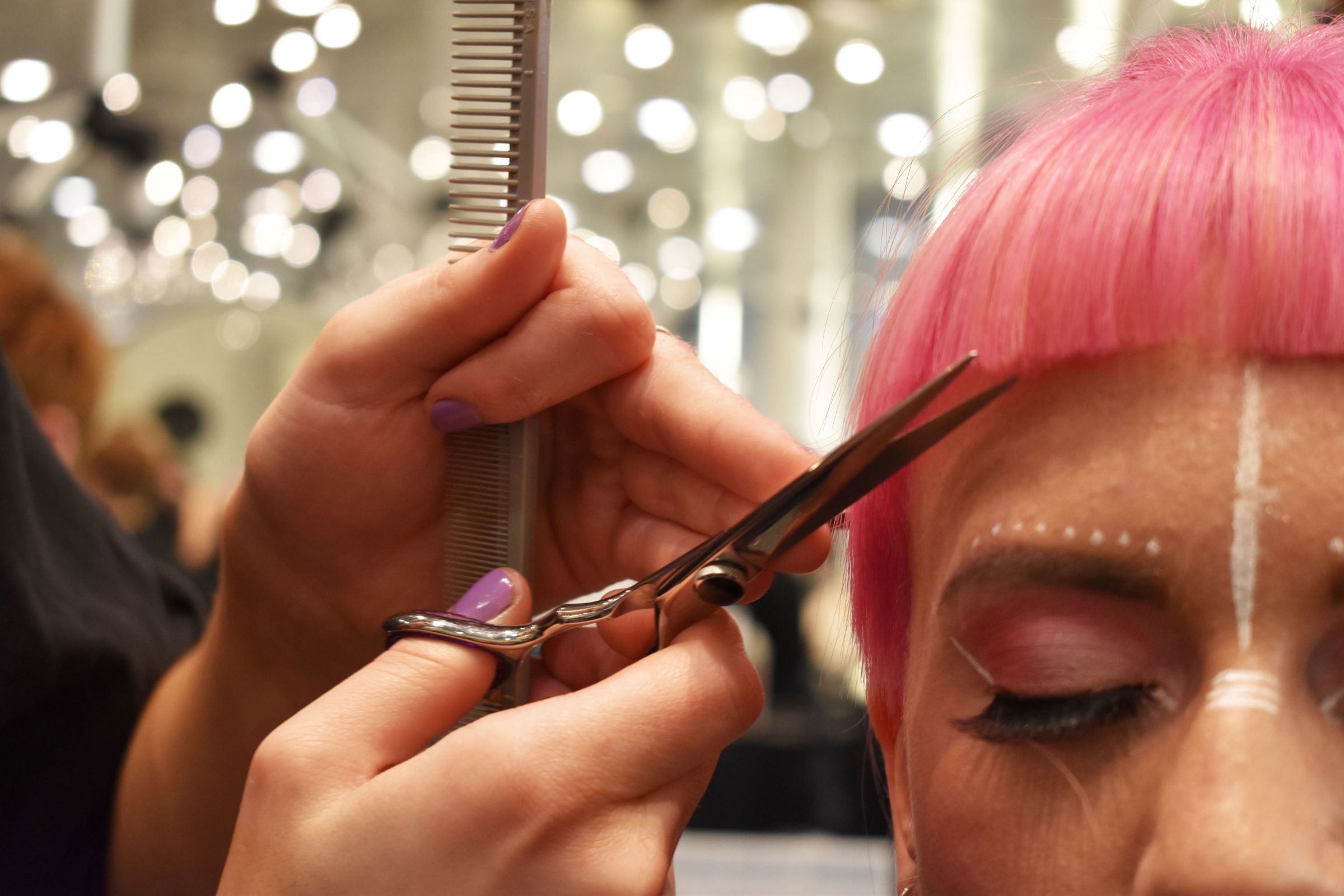 Frisørelev klipper pandehår på en model, som har fået farvet håret pink og er blevet sminket i ansigtet med hvide prikker og streger