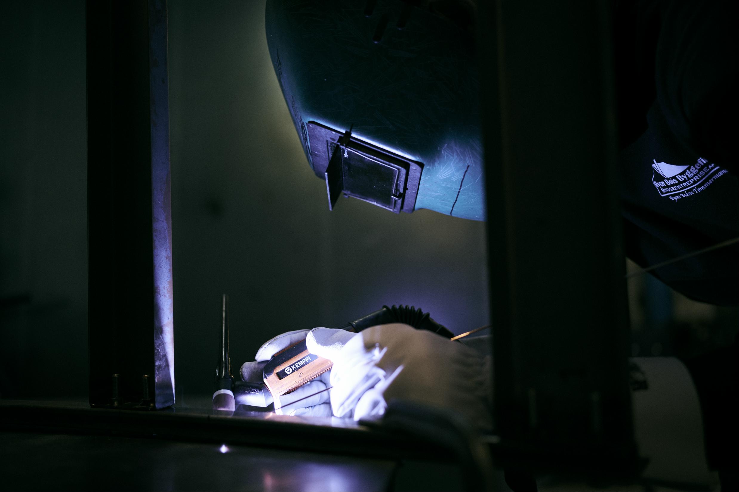 En lærling på skolens smedeværksted, med ansigtet gemt bag svejsehjelmen, er i færd med at svejse.