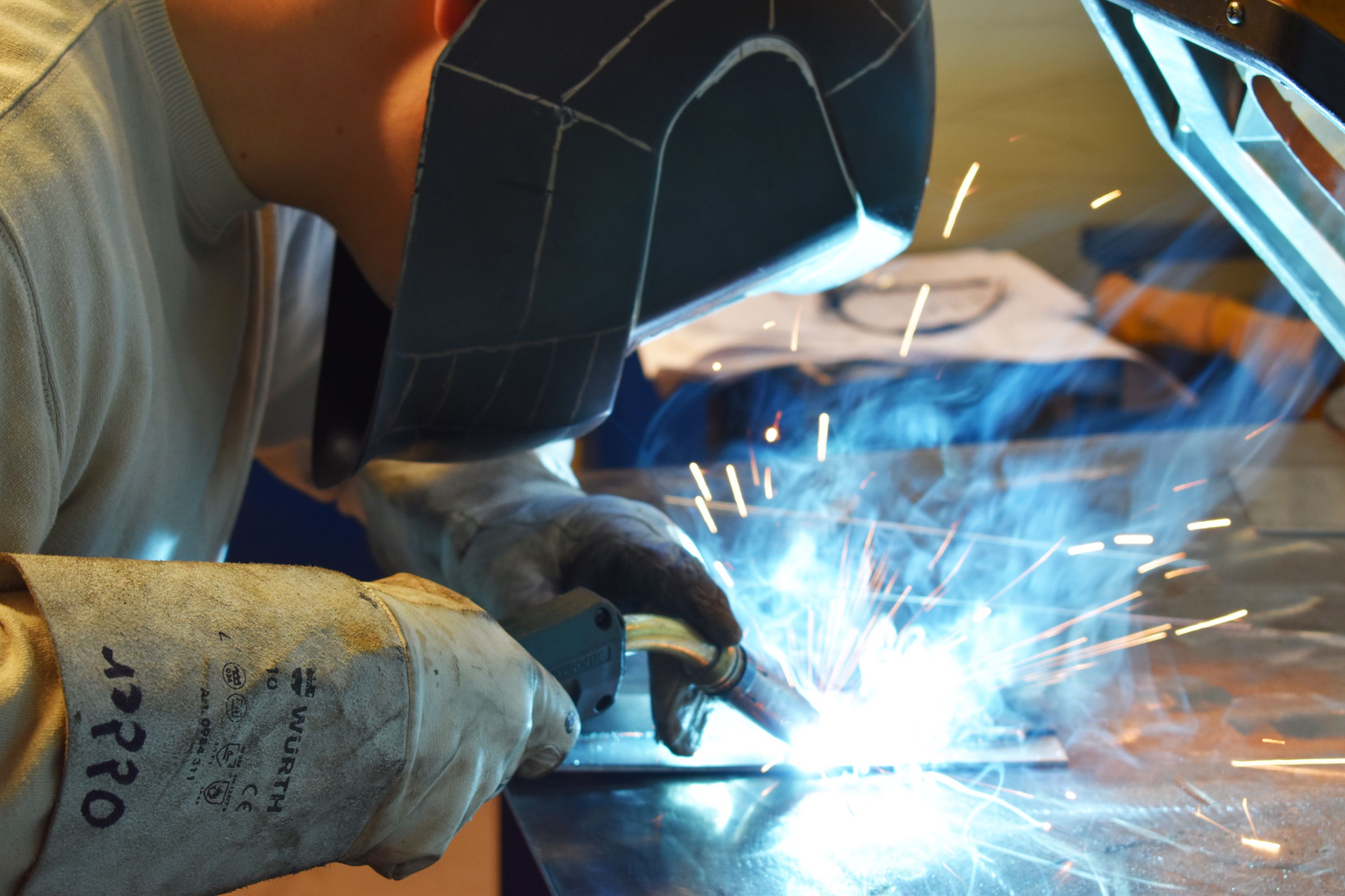 En smedelærling iført svejsehjelm og beskyttelseshandsker svejser, så der kommer blå flammer og gnister