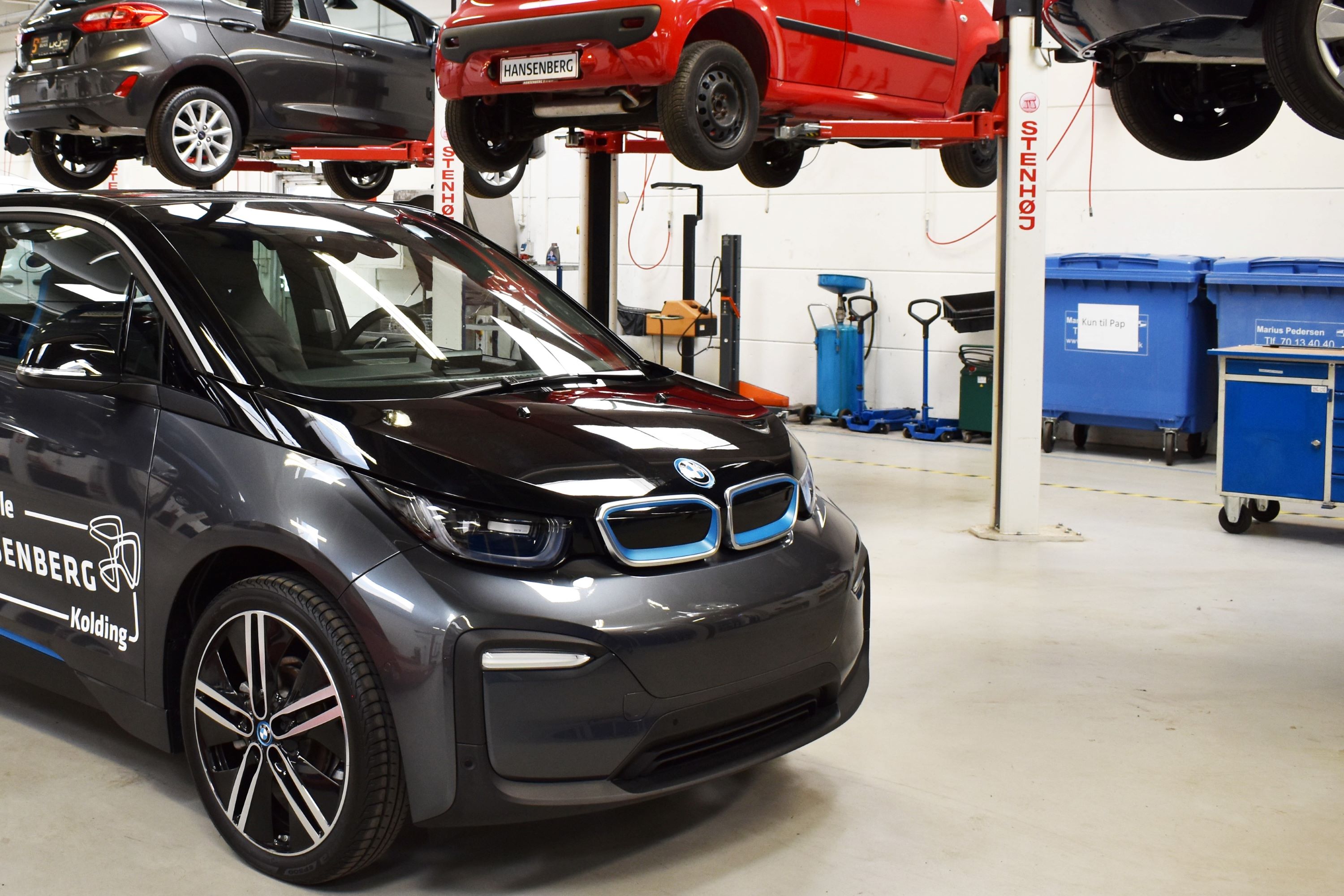 Elbilen er en af de biler, som mekanikereleverne øver sig på, og bagved hænger andre biler på nogle af de mange autolifte på skolens værksted.