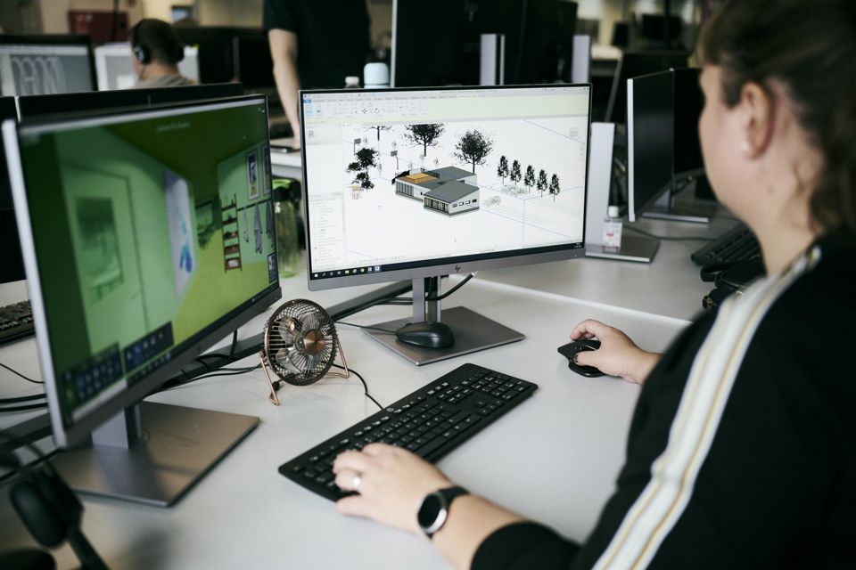 En teknisk designerelev er ved at designe en bygning og tjekker bygningen i forskellige udgaver af 3D på de to skærme.